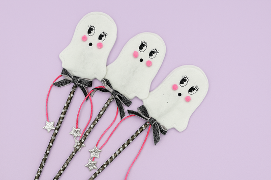 Three ghost-shaped  party wands with black bows on a light purple background