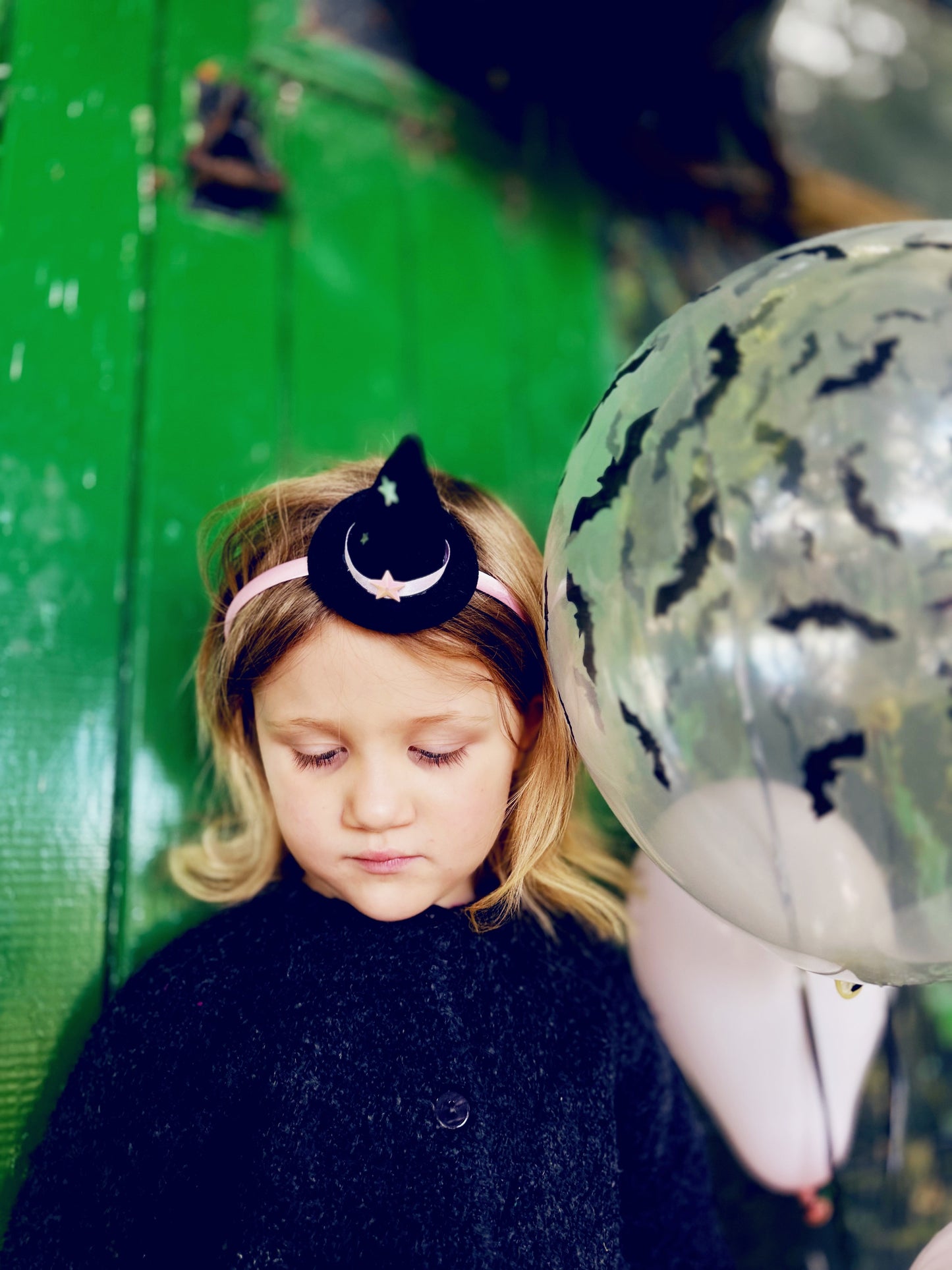 Child wearing a headband with a moon and star design, standing next to a green wall and balloons.