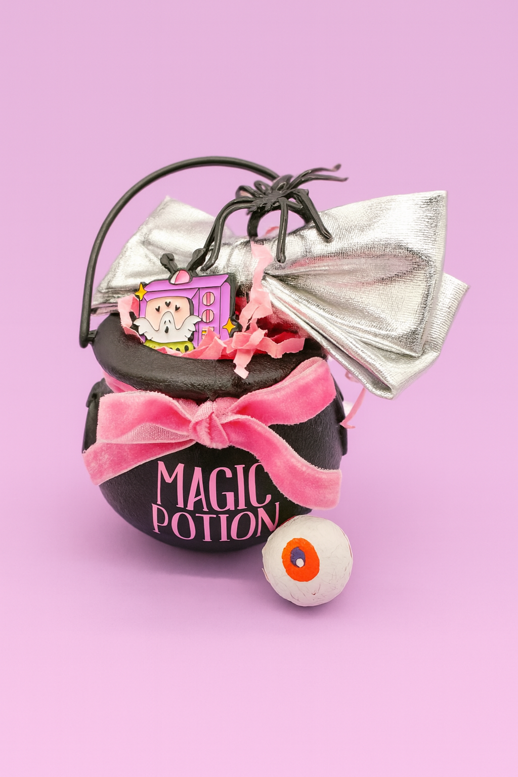 Decorative Halloween cauldron labeled 'Magic Potion' with spider, money, and candy on a pink background