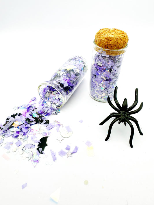 Halloween-themed confetti in a glass bottle with a cork lid, surrounded by spider decorations on a white background.