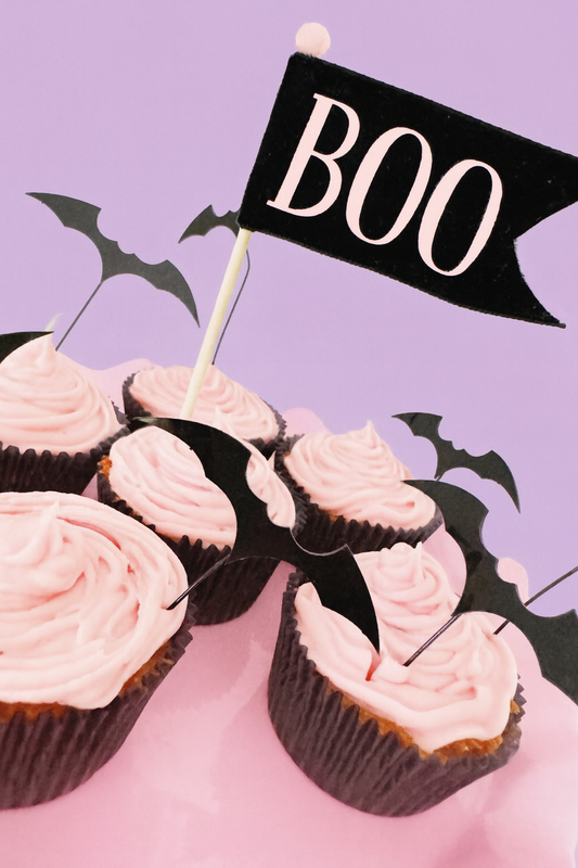 Cupcakes with pink frosting and black bat decorations on a purple background with a 'BOO' flag.