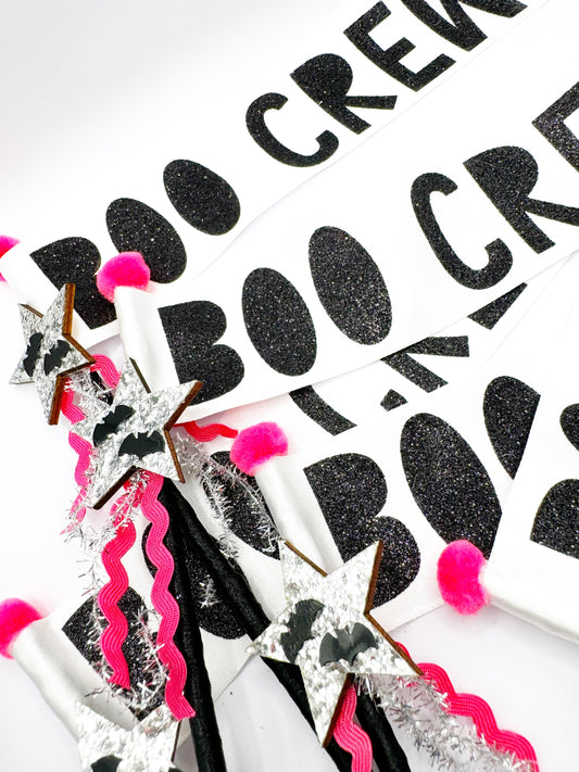 Decorative halloween boo crew wands with black glittery text, pink pom-poms, and decorative ribbons on a white background.