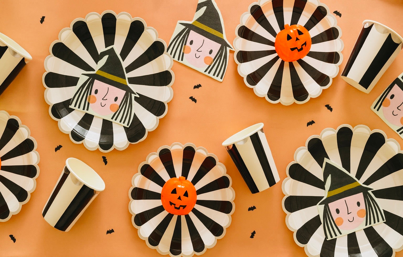 Halloween-themed paper plates and cups with black, white, and orange designs on a peach background.