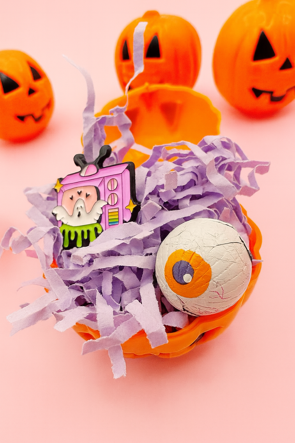 Halloween-themed product with purple ribbon, small house figure, and eyeball on a pink background with pumpkins.